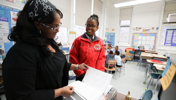 A city year corps member with a teacher in a classroom with students in a circle in the back ground