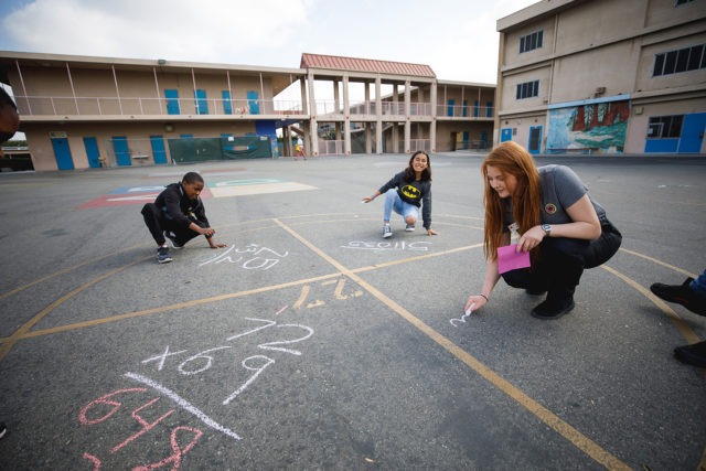 city year americorps member and student drawing out math problems with chalk on the black pavement in a school courtyard