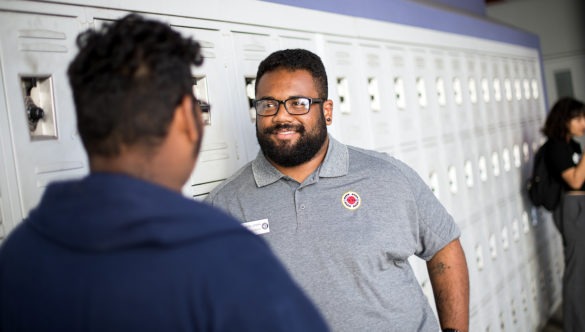 city year americorps member talking with a high school student as they stand in front of lockers in a school hallway