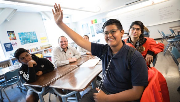 A student raising his hand as he sits with other students and city year americorps members in a classroom